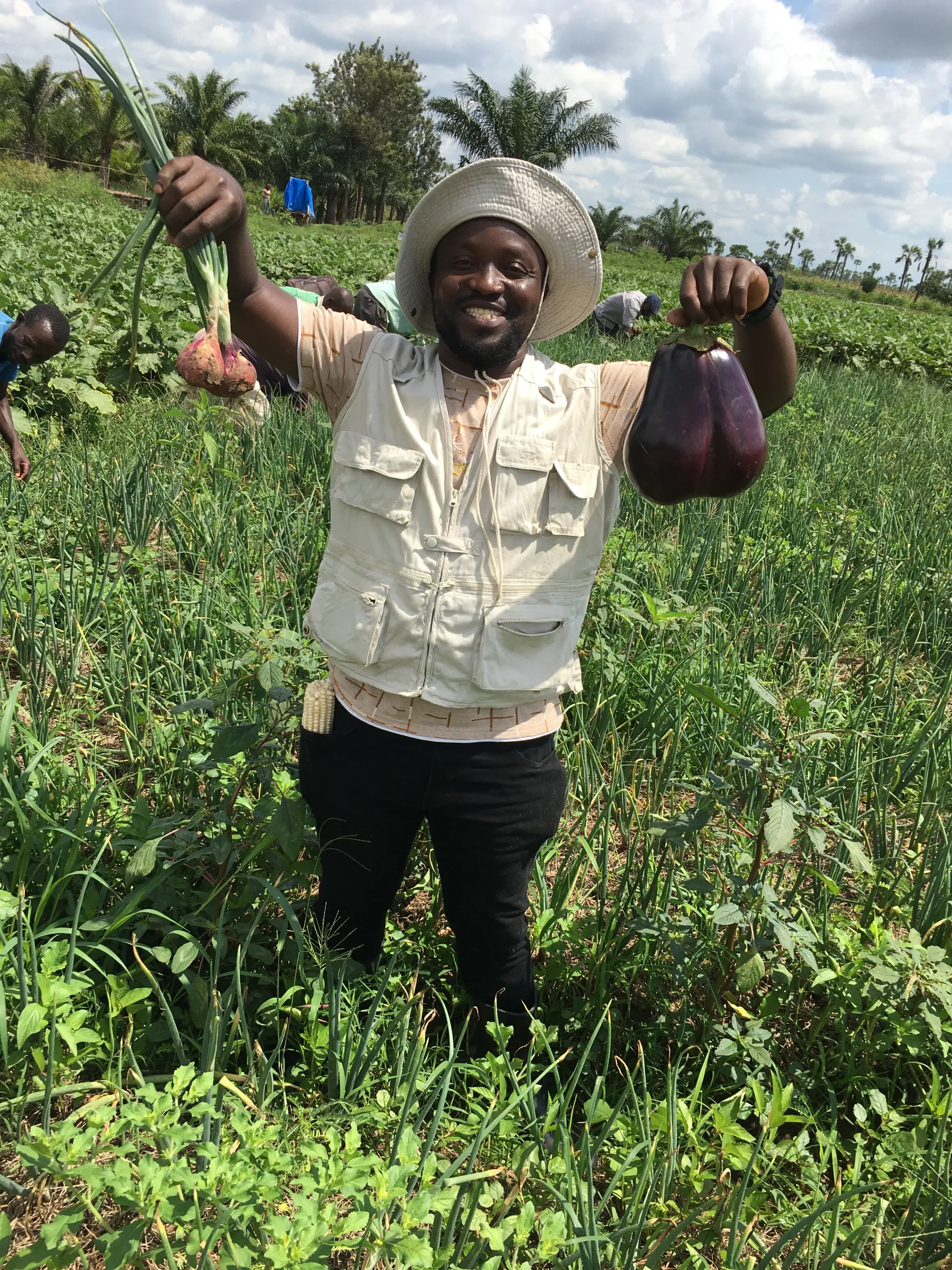 A BORA Alliance trainer in a field vest holding up freshly harvested onions and aubergines in a nutrition garden